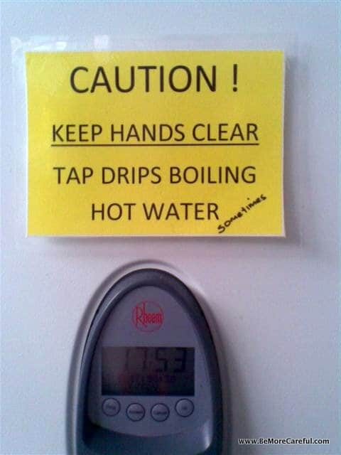 A yellow caution sign reads "CAUTION! KEEP HANDS CLEAR TAP DRIPS BOILING HOT WATER" with a handwritten note "sometimes" added at the bottom. Below the sign is a digital temperature display showing "115.3" and the Rheem logo.






