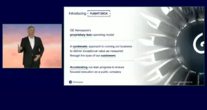 **Alt Text:** A presentation slide from GE Aerospace's Investor Day featuring CEO Larry Culp. The slide is titled "Introducing - FLIGHT DECK" and includes the following text: - GE Aerospace's **proprietary lean operating model** - A **systematic** approach to running our business to deliver exceptional value as measured through the eyes of our **customers** - **Accelerating** our lean progress to ensure focused execution as a public company Larry Culp is pictured on the left side of the slide, wearing a black suit and speaking against a backdrop with a gradient of warm colors. On the right side, there is an image of a turbine or engine component. The GE Aerospace logo is displayed at the bottom, and the slide is marked "2024 GE Aerospace Investor Day" with the page number 13.