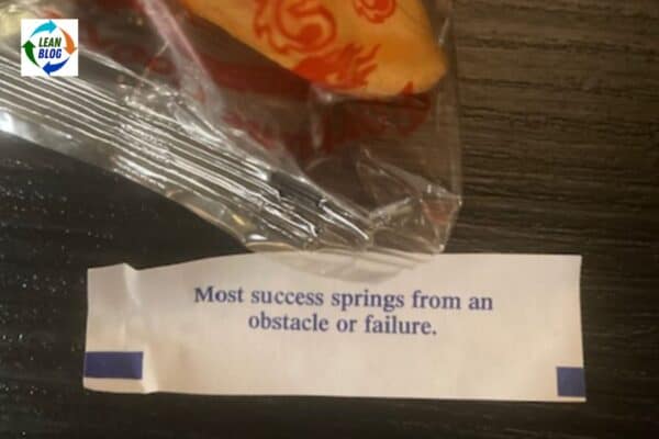 A fortune cookie partially wrapped in clear plastic packaging rests on a dark surface. Next to it, the fortune reads, "Most success springs from an obstacle or failure."